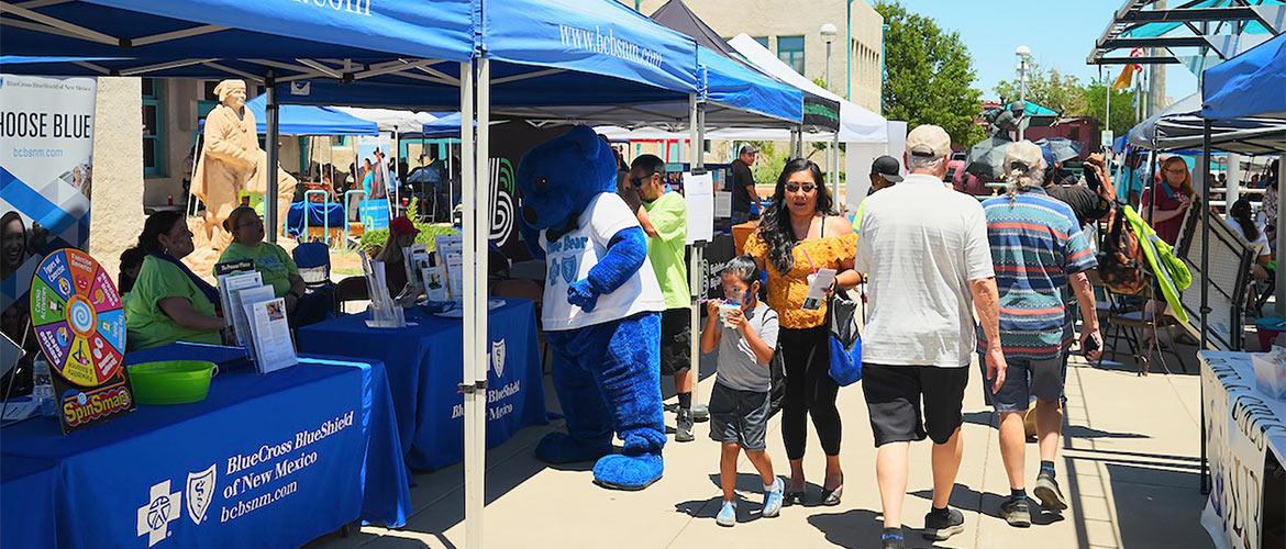A woman and child walk by a Blue Cross and Blue Shield of New Mexico information tent and its Blue Bear mascot at an outdoor health fair