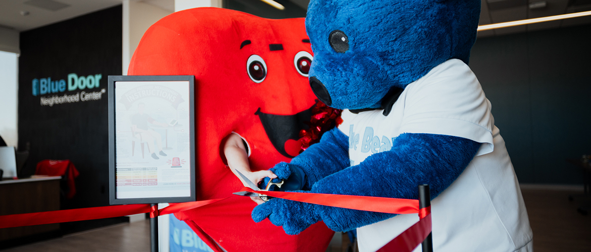 Person dress in large red heart costume cuts red ribbon at Blue Door Neighborhood Center with person in blue bear costume