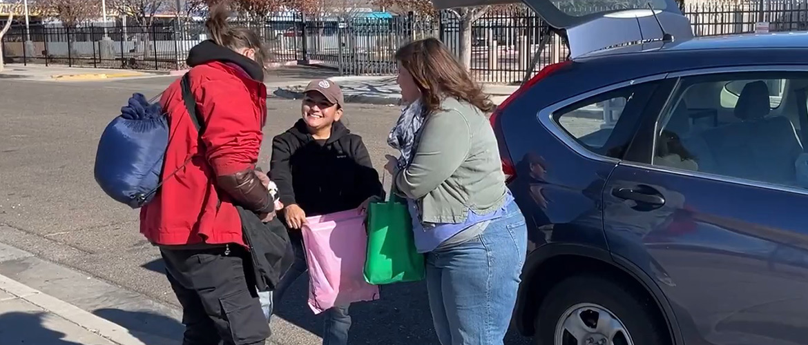 Two women gather around a woman holding open a bag as she stands near the back of an SUV