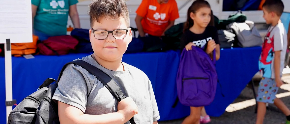 A boy with glasses holds a new backpack in front of a Blue Cross and Blue Shield of New Mexico Care Van
