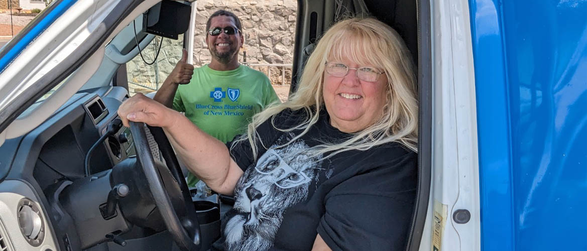 Una mujer sentada en el asiento del conductor de una camioneta mientras un hombre muy sonriente saluda por la puerta del acompañante detrás de ella.