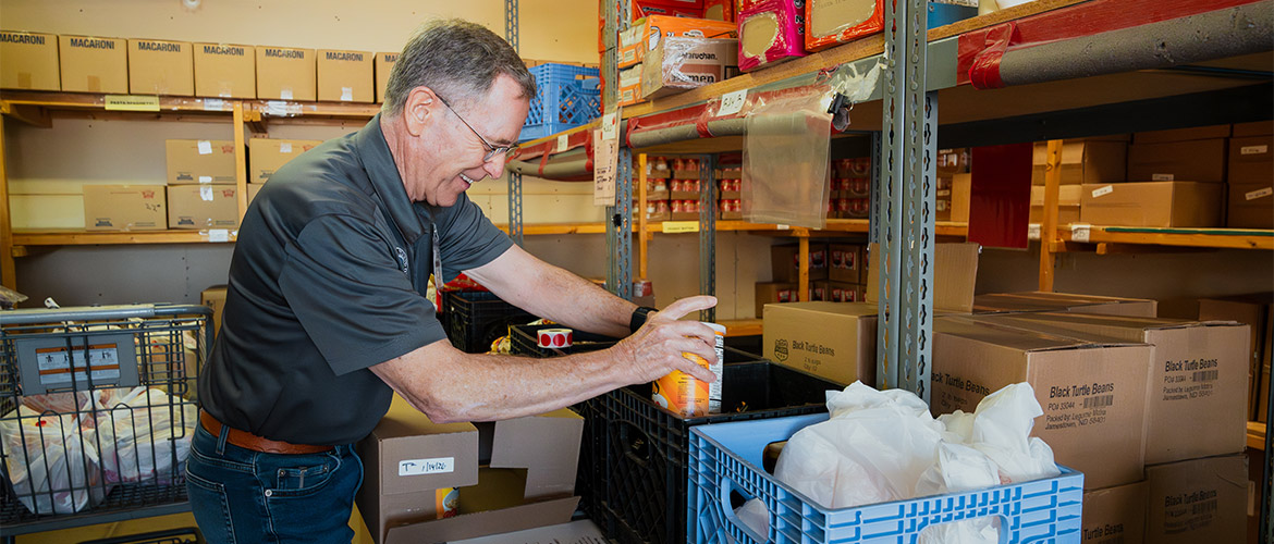 An older man in jeans and a tucked in polo shirt stocks grocery items at a food pantry