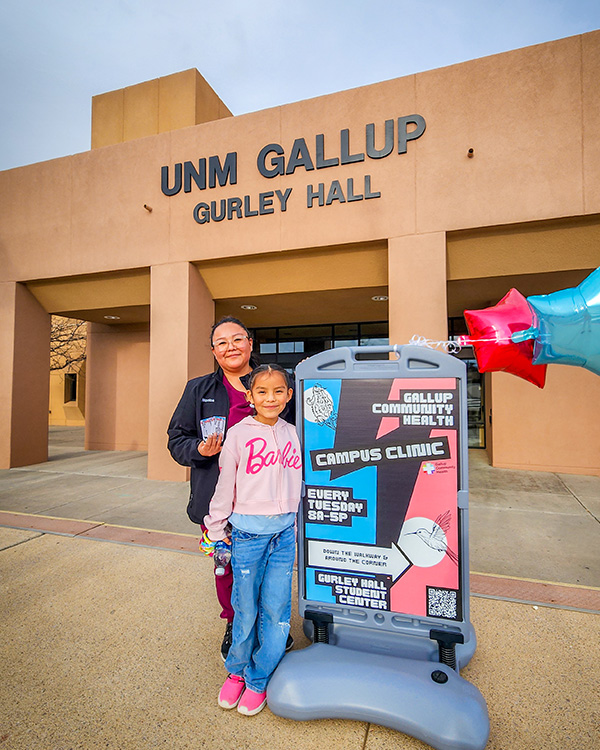 A woman and child stand outside a school building next to a sign announcing a campus clinic. 