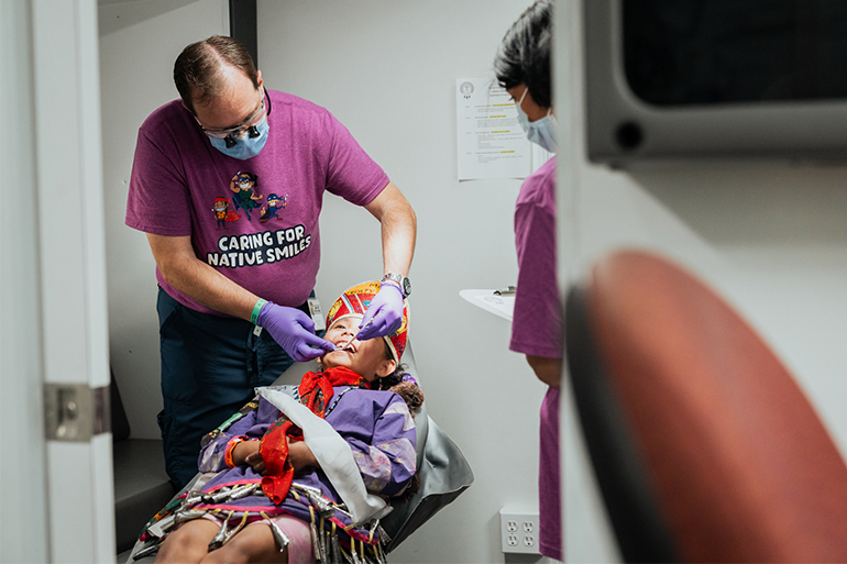 A dentist in a purple shirt checks the teeth of little girl laying down in chair