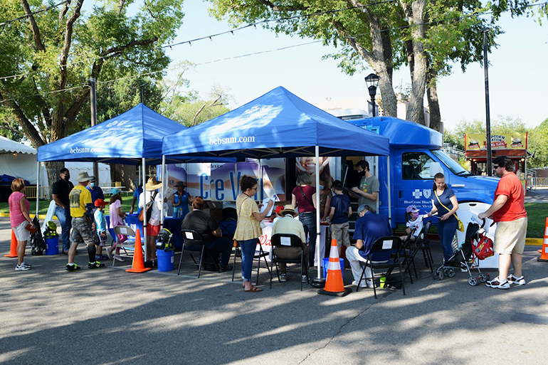 People standing and sitting in fold-up chairs under blue tents outside near Care Vans