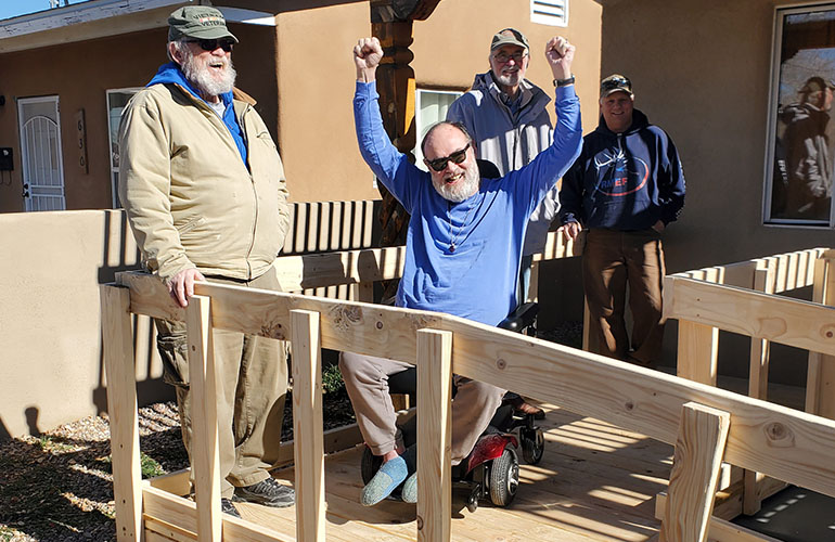 A man in a wheelchair on a ramp in front of his house holds his hands in the air in celebration