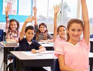 Middle school aged children seated at their desks in a classroom with their hands raised