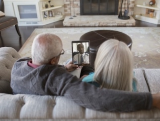 An elderly couple sitting on a couch with their backs to the camera, looking at their doctor on a mobile tablet