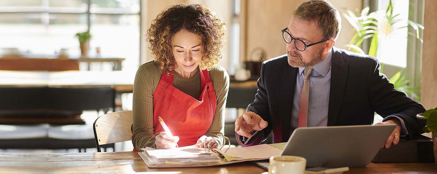 A woman in an apron writes on paperwork while a man in a suit assists