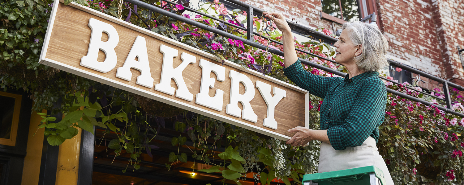 A woman standing on a ladder outside of her small business hangs a sign that says "BAKERY"