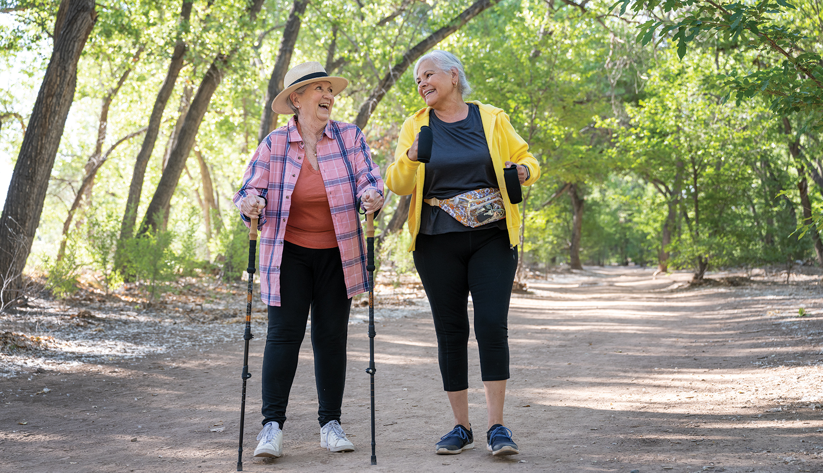 Two smiling senior women walking on a treelined trail in a New Mexico park.